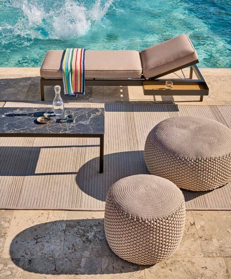 Eine Szene am Pool mit einem beigen Loungesessel mit einem bunt gestreiften Handtuch, zwei WEISHÄUPL Weishäupl Pouf Hocker Medium – Indoor – Outdoor in Taupe (55 cm) und einem Couchtisch mit Marmorplatte mit einer Karaffe und Gläsern. Klares blaues Wasser spritzt aus dem Pool und sorgt sowohl drinnen als auch draußen für ein Gefühl der Entspannung und sommerlichen Freizeit.