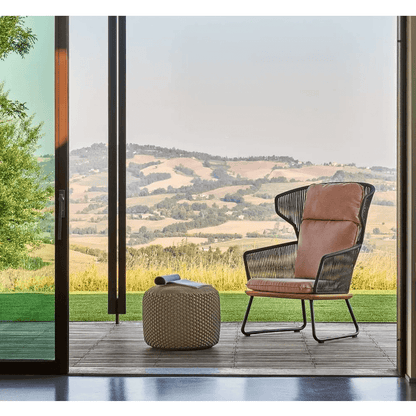 Ein moderner Stuhl mit taupefarbenem Kissen und ein WEISHÄUPL Pouf Hocker Medium – Indoor – Outdoor, Taupe 55 cm stehen auf einer Holzterrasse mit Blick auf eine malerische Landschaft mit sanften Hügeln. Die Innen-/Außenumgebung wird von einer großen offenen Glasschiebetür eingerahmt, die die Räume nahtlos miteinander verbindet.